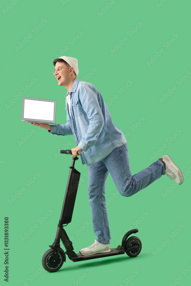 Young man with blank laptop riding kick scooter on green background