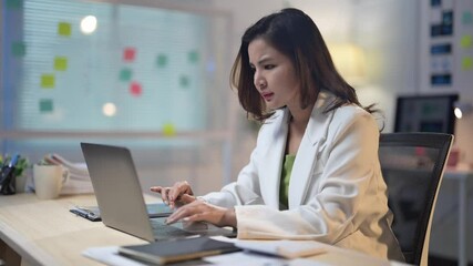 Wall Mural - A woman is sitting at a desk with a laptop in front of her. She is wearing a white jacket and she is working on her computer