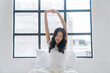 © kaew6566 - Healthy asian woman waking up relax muscles beside window early in morning. Happy young girl stretching raised arms muscles body cozy after waking up sitting on bed with pillow at apartment. Lifestyle