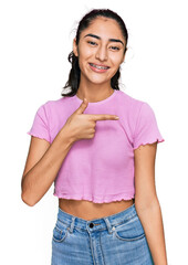 Wall Mural - Hispanic teenager girl with dental braces wearing casual clothes cheerful with a smile on face pointing with hand and finger up to the side with happy and natural expression