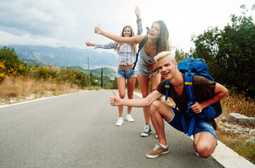 Wall Mural - cheerful interracial friends standing on road showing hitchhiking gestures. tourism traveling