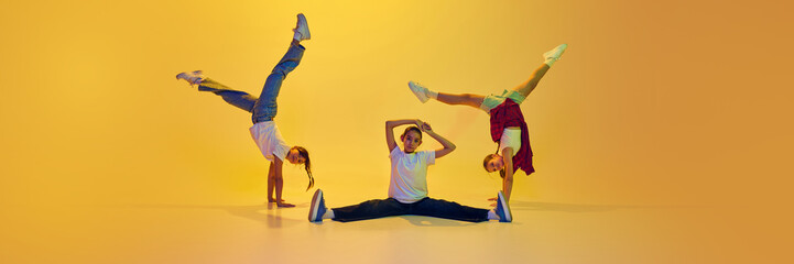 Dynamic image of three active teen girls in motion, wearing casual clothes, dancing hip hop against yellow background in neon light. Concept of modern dance, youth, active lifestyle, hobby