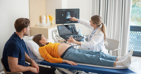 Aufkleber - Young Pregnant Woman Lies on a Clinic Bed while a Caucasian Female Obstetrician Performs an Ultrasound Procedure. Her Partner Sits Nearby, Taking Her Hand, Watching the Screen with Joyful Anticipation