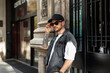 © alones - Fashion handsome man with black cap in fashionable denim clothes with denim vest puts on sunglasses and stands near building in Barcelona