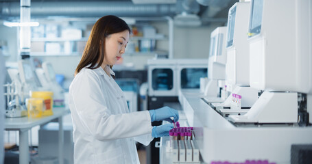 Leinwandbilder - Young Asian Scientist Carefully Prepares Test Vials for Genetic Analysis as Her Male and Female Colleagues Prepare Medical Equipment in the Background