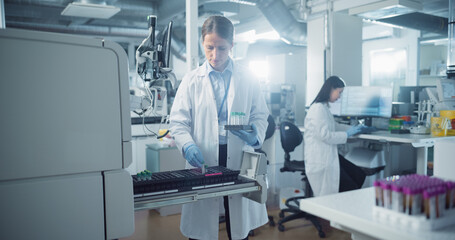 Leinwandbilder - Female Scientist Carefully Loads Test Tubes into a Machine for Genetic Analysis as Her Asian Coworker Prepares Equipment on a Computer in the Background in a Modern and Sterile Laboratory