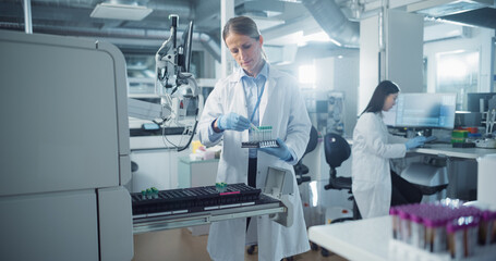 Leinwandbilder - Female Scientist Carefully Loads Test Tubes into a Machine for Genetic Analysis as Her Asian Coworker Prepares Equipment on a Computer in the Background in a Modern and Sterile Laboratory