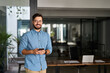© insta_photos - Happy professional Latin young business man looking at camera at work, portrait. Confident smiling businessman leader, male executive manager entrepreneur standing in office with smartphone in hands.