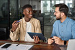 © insta_photos - Two busy diverse male employees using digital tablet talking in office. Professional young business men workers team having conversation working on project sharing ideas sitting at work desk.