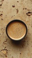 Poster - A top view captures a simple black cup filled with rich, frothy coffee, resting on a textured beige surface. This serene setting invites moments of relaxation and contemplation