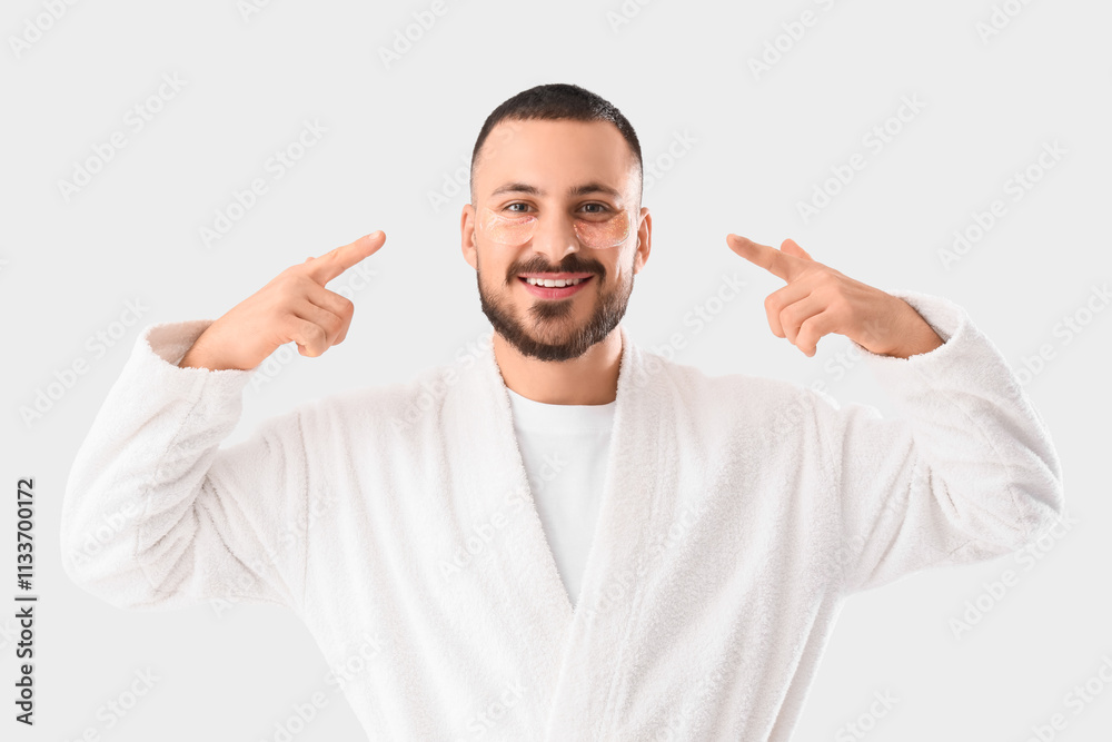 Smiling young man in robe pointing to eye patches on his face on white background