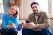 © Raquel - A cheerful young couple sitting indoors, both holding smartphones and smiling warmly. The woman leans slightly, creating a relaxed and friendly atmosphere in a cozy setting.