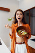 © SHOTPRIME STUDIO - Surprised woman in orange shirt enjoying fresh salad, healthy eating concept with vibrant colors in a modern kitchen
