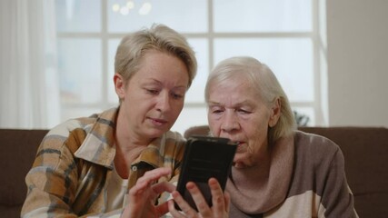 Two women elderly mother and middle aged daughter using smartphone while sitting on sofa in room.