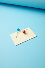 Aufkleber - A group of a pin and a thumbtack sit on a small piece of blank notepaper on a blue desk surface._00001_