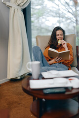 Canvas Print - Young woman enjoying a snack while reading a book, lounging in a cozy armchair, vibrant earthy tones, peaceful ambiance, indoor setting, autumn vibe, comfort lifestyle