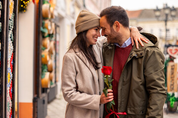 Mid adult Caucasian couple indulges in romantic window shopping during Valentine's Day. The woman holds a rose and wears a cozy coat, while the man sports a warm jacket.