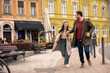 © Dorde - Caucasian couple joyfully running on a charming street. The man wears a green jacket and red sweater, the woman in a beige coat and hat.