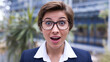 © Tom - Surprised young Caucasian woman with short hair and glasses, dressed in a business suit, expressing excitement outdoors amidst greenery.