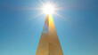 © Thiago - A tall golden obelisk reaching into a clear blue sky with the sun shining brightly above, creating a striking silhouette. Obelisk. Illustration