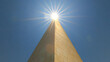 © Thiago - A tall golden obelisk reaching into a clear blue sky with the sun shining brightly above, creating a striking silhouette. Obelisk. Illustration