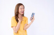 © suphaporn - Cheerful and Beautiful Asian Young Woman in Yellow T-Shirt Laughing While Looking at Smartphone, Expressing Happiness and Excitement, Isolated on White Background, Concept of Digital Communication