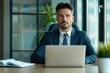 © Vilaysack - A professional man in a suit sits at a desk with a laptop, looking thoughtfully at the camera, surrounded by a modern office setting.