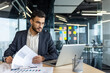 © Liubomir - Mature man businessman sits at a desk, analyzing reports and working on a laptop. The professionally dressed individual is based in a bright, modern office with an organized workspace.