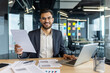 © Liubomir - Mature male businessman examining documents and using a laptop at his desk in a modern office. He appears confident and professional while focusing on work.