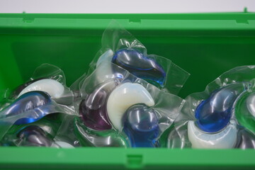 Things, objects for daily use, cleansing. Round polyethylene capsules filled with white, blue, green, purple laundry detergent, clothes located in a green plastic container on white plastic backgroud.