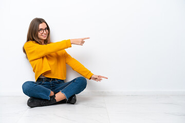 Wall Mural - Young caucasian woman sitting on the floor isolated on white background pointing finger to the side and presenting a product