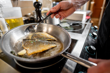 Wall Mural - A chef is frying fish in a stainless steel pan, using a spoon to baste it with oil and herbs. The kitchen setting includes olive oil and seasonings, reflecting a culinary vibe.