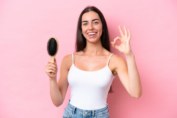 Wall Mural - Young caucasian woman with hair comb isolated on pink background showing ok sign with fingers