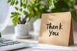 © ольга  - A brown postcard with the words 'Thank you' on the table next to a laptop and a potted plant.