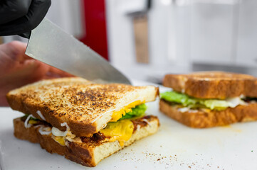 Wall Mural - A close-up of a hand slicing a toasted sandwich with melted cheese, lettuce, and mayonnaise. A second sandwich sits nearby, showcasing a delicious, golden-brown crust in a kitchen setting.