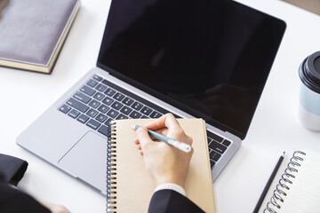 Poster - Detailed view of a woman's hand jotting down notes in a notepad on a modern laptop, with a blurred background