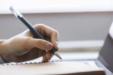 Poster - The image focuses on a woman's hand scribbling in a notepad on a stylish laptop, set against a blurry scene
