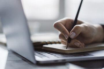 Poster - A woman's hand is seen in close detail, writing in a notepad on a sleek laptop, background blurred