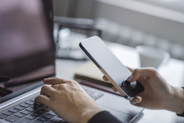 Wall Mural - Woman working intently on her advanced smartphone at her desk, laptop open and ready nearby set against the indistinct buzz of the office environment