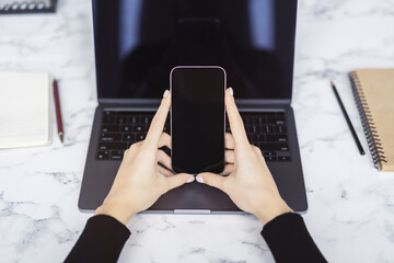 Wall Mural - Woman working intently on her advanced smartphone at her desk, laptop open and ready nearby set against the indistinct buzz of the office environment