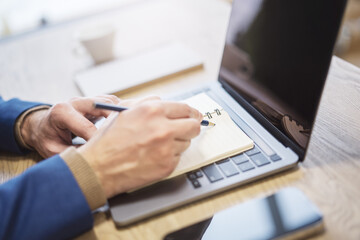 Poster - Close perspective of a man's hand making entries in a notepad on a cutting-edge laptop, with a soft blur in the surrounding