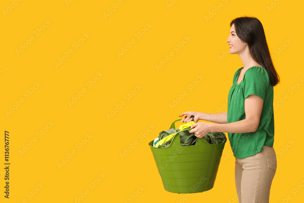 Young woman holding basket with clean laundry on yellow background