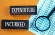 © Zhanna - EXPENDITURE INCURRED - words on brown wooden bars against the background of a magnifying glass and a table with numbers