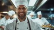 © svastix - A cheerful head chef in a pristine white uniform and hat stands smiling in a bustling kitchen surrounded by a collaborative cooking team, reflecting teamwork and culinary passion.