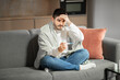 © Prostock-studio - A young European man in eyeglasses sits on a couch at home, wrapped in a blanket. He appears concerned as he checks his temperature with a thermometer, showing signs of illness.