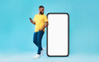 © Prostock-studio - Cheery millennial black guy with mobile device looking at giant smartphone on blue background, mockup for mobile app on white screen. Cellphone website template, cool online store ad
