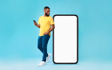 Poster - Cheery millennial black guy with mobile device looking at giant smartphone on blue background, mockup for mobile app on white screen. Cellphone website template, cool online store ad