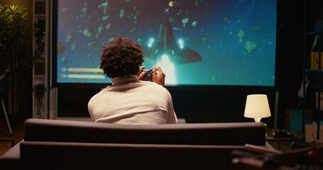 African american gamer playing classic arcade space shooter videogame on big smart TV, shooting asteroids. Zoom in shot on man using gaming console to solve missions in singleplayer game