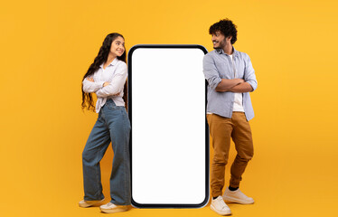 Poster - Mobile offer. Millennial indian couple leaning on giant smartphone with mockup, advertising or promoting app or website over yellow studio background