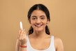 © Prostock-studio - A young Indian woman is happily showcasing a skincare product while pampering herself. She exudes confidence and beauty against a warm, neutral backdrop that highlights her glowing skin.